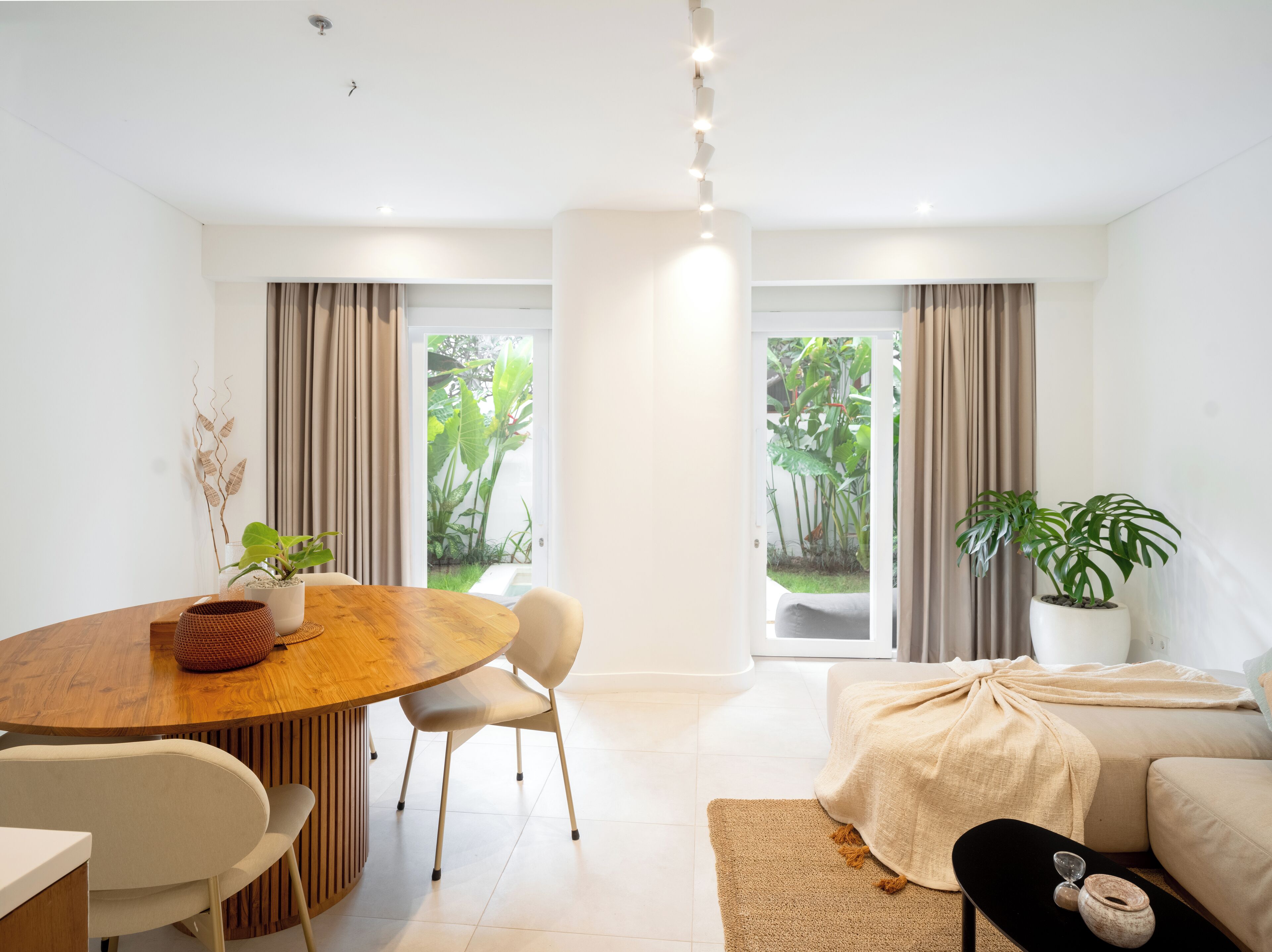 A modern living room with a wooden dining table, two chairs, a sofa, and a large window with curtains.