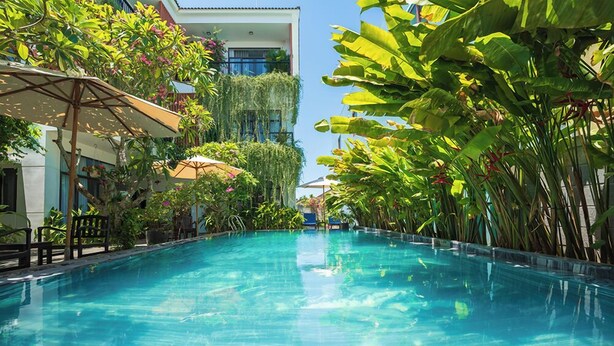 A swimming pool with clear blue water, surrounded by lush greenery and a building with balconies. - Coco Island Villa (Da Nang)