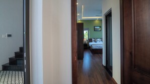 A hallway leading to a bedroom with a bed, a nightstand, and a window with a view of greenery. - Coco Island Villa (Da Nang)