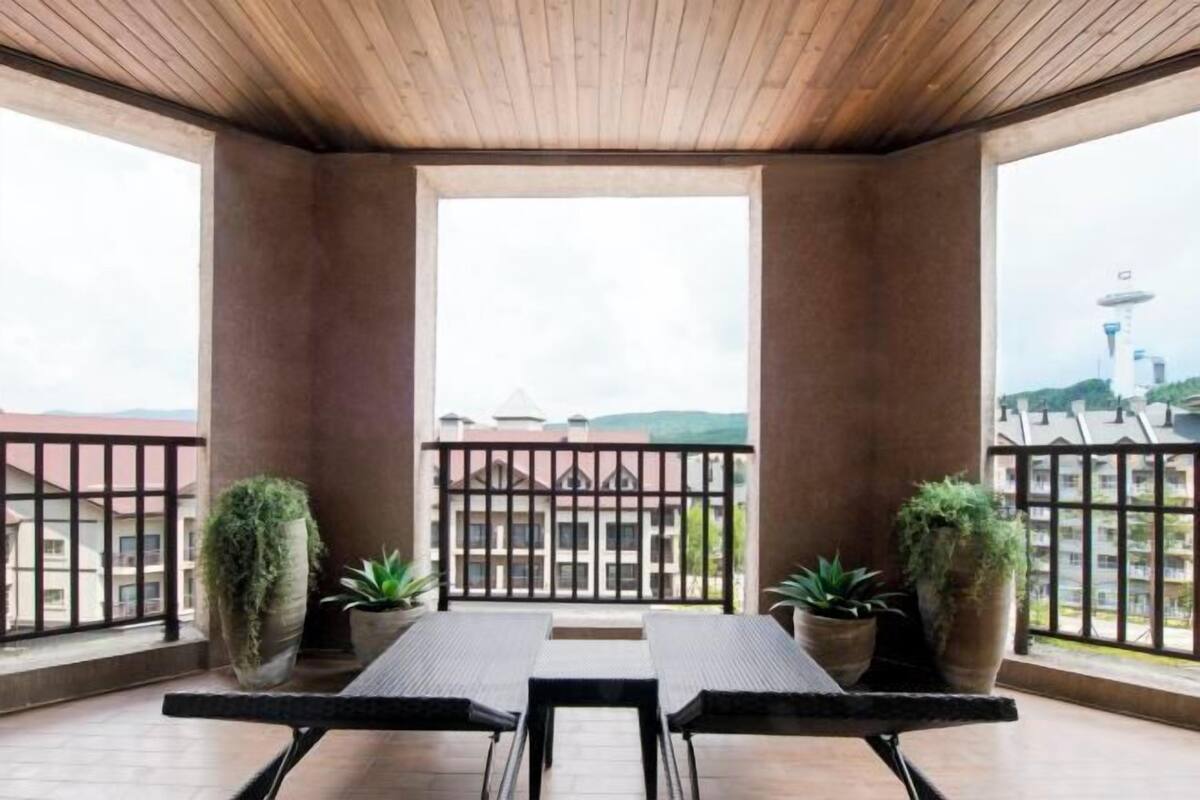 Een balkon met een houten plafond, twee planten in potten, een tafel en stoelen.