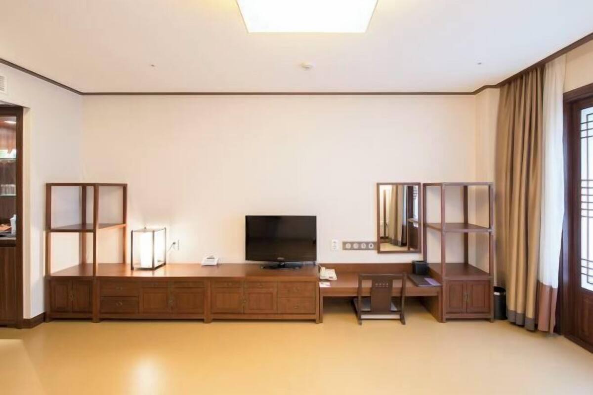 Een hotelkamer met een grote flatscreen televisie, een bureau met een stoel, een boekenplank en een raam met gordijnen.