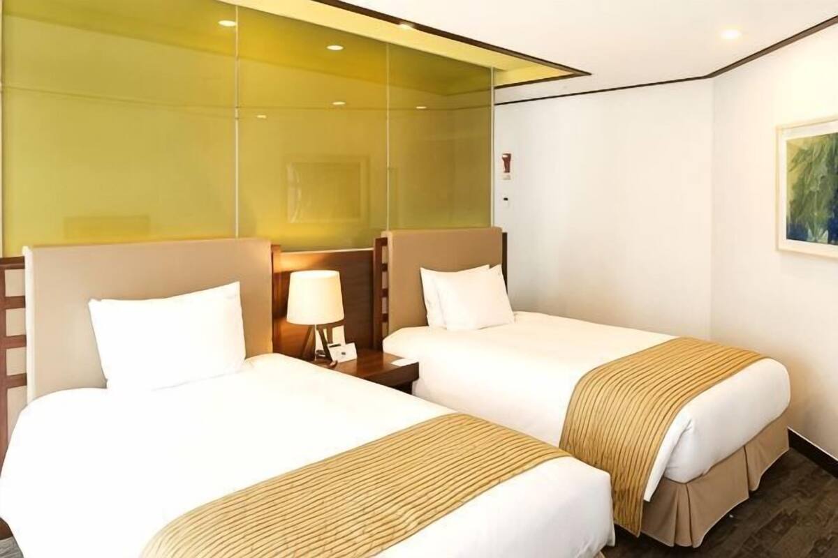 Een hotelkamer met twee bedden, een glazen scheidingswand, een nachtkastje met een lamp en een schilderij aan de muur.