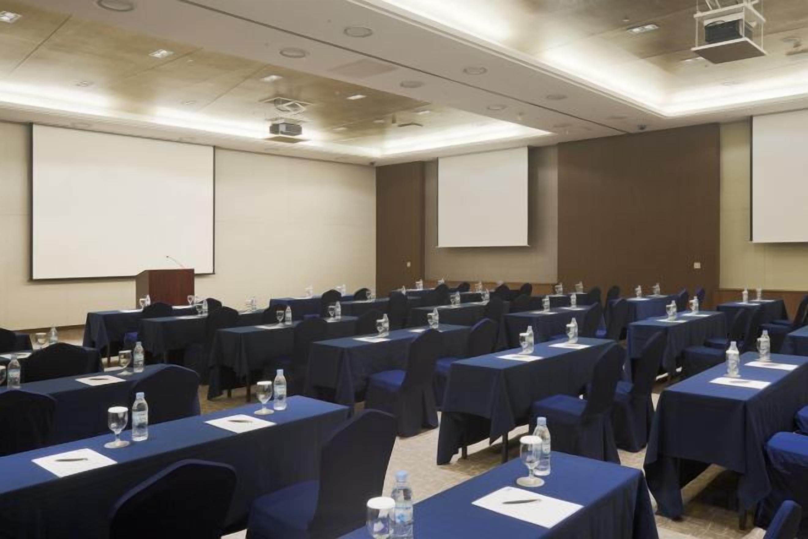 Sala de conferências com mesas, cadeiras e telas de apresentação dispostas em fileiras.