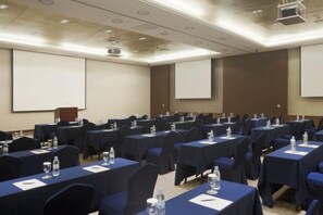Un salón de conferencias con filas de mesas, sillas y pantallas para presentaciones.