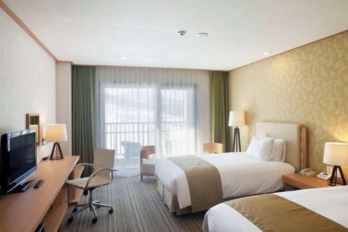 Een hotelkamer met twee bedden, een bureau met een televisie, een stoel en een raam met dunne gordijnen.