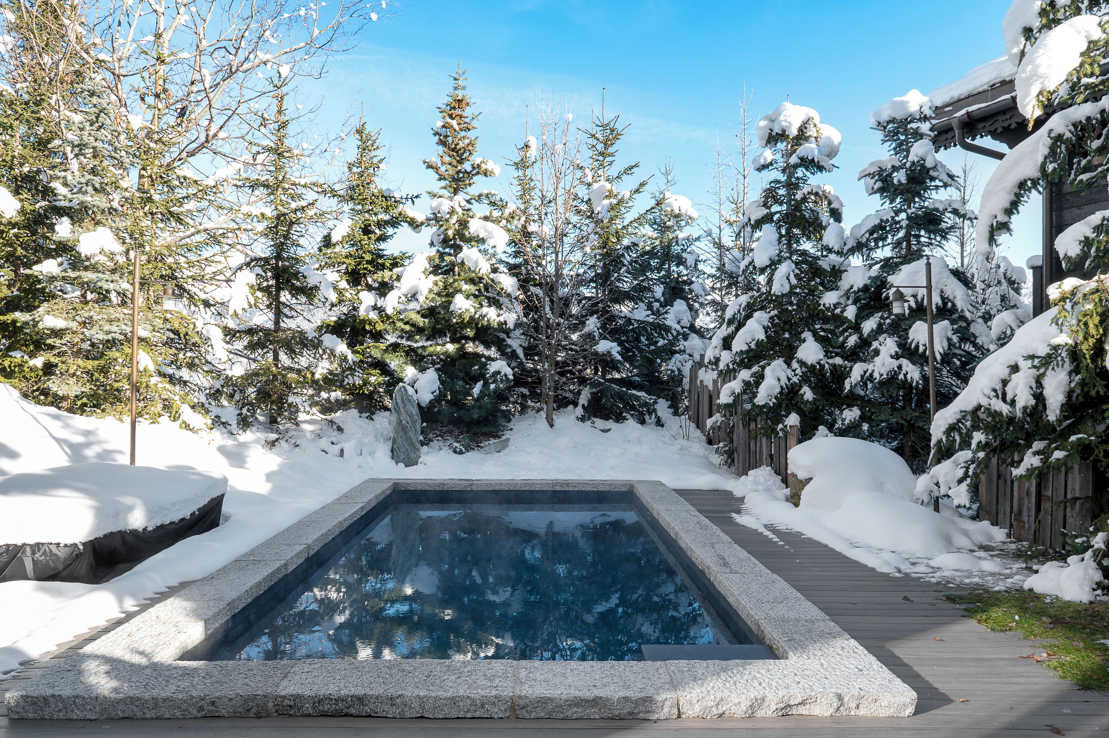 Una piscina rectangular al aire libre rodeada de árboles cubiertos de nieve y una terraza de madera.