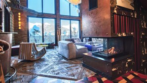 A spacious living area with large windows, a fireplace, and a unique wooden floor design. - Le Hameau de Marcandou (Courchevel)