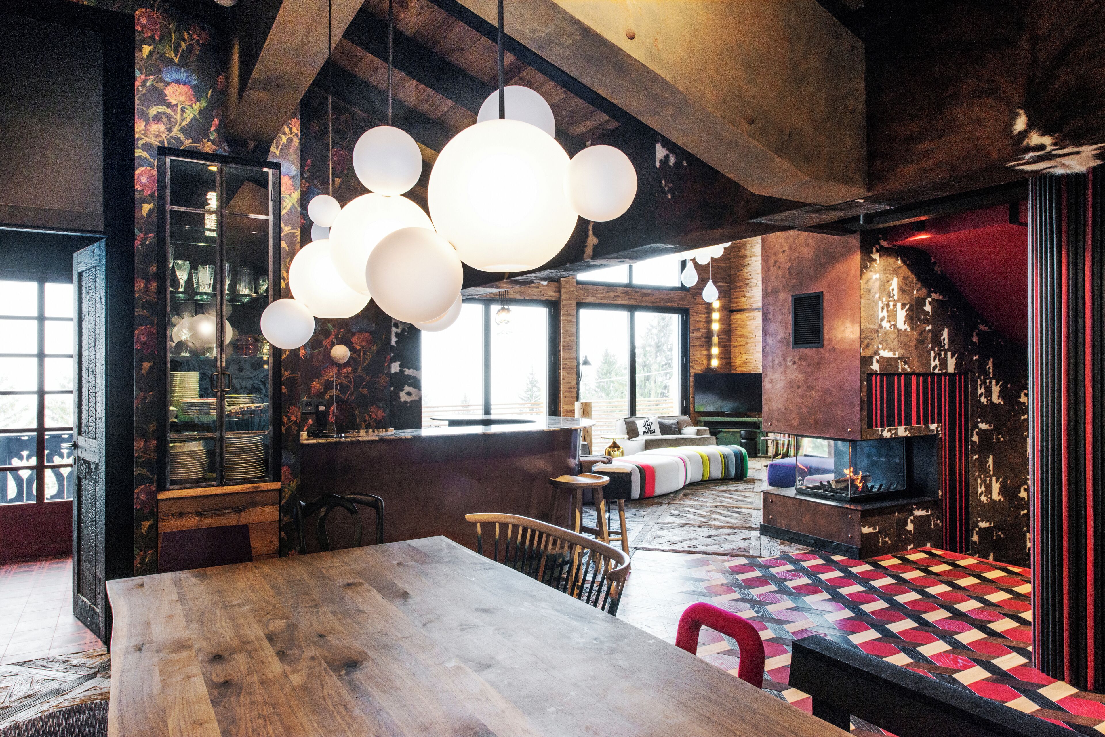 Uma sala de estar ampla com uma grande mesa de madeira, um sofá e um tapete colorido com padrão. Há também um espaço de bar com banquetas e uma televisão.