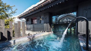 A clear pool with a wooden deck, a wooden fence, and a building with a red door and a snow-covered roof. - Le Hameau de Marcandou (Courchevel)