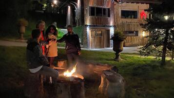Un groupe de personnes est réuni autour d’un feu de camp dans un jardin, avec une maison en bois en arrière-plan.
