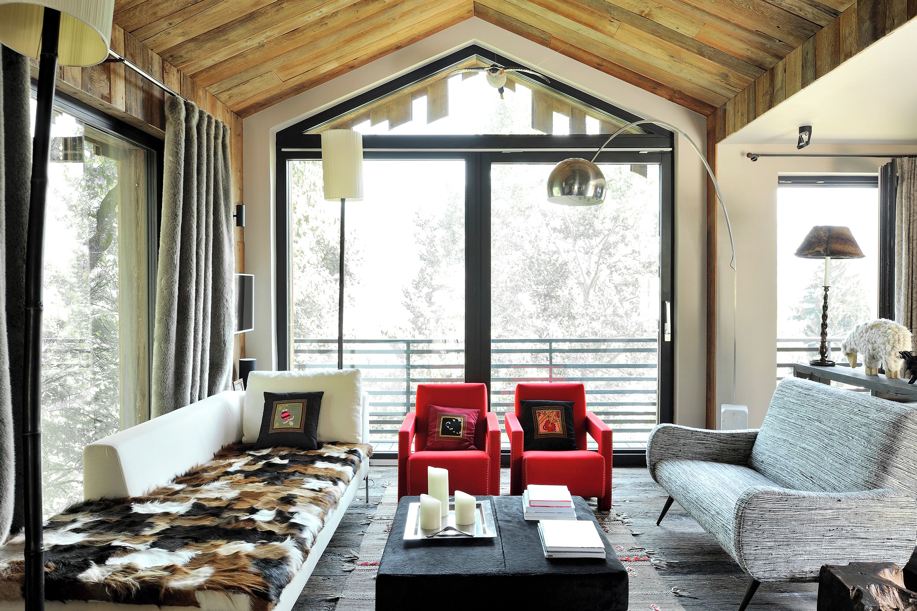 A cozy living room with a high ceiling, large windows, and a mix of seating including a sofa, two red chairs, and a grey couch.