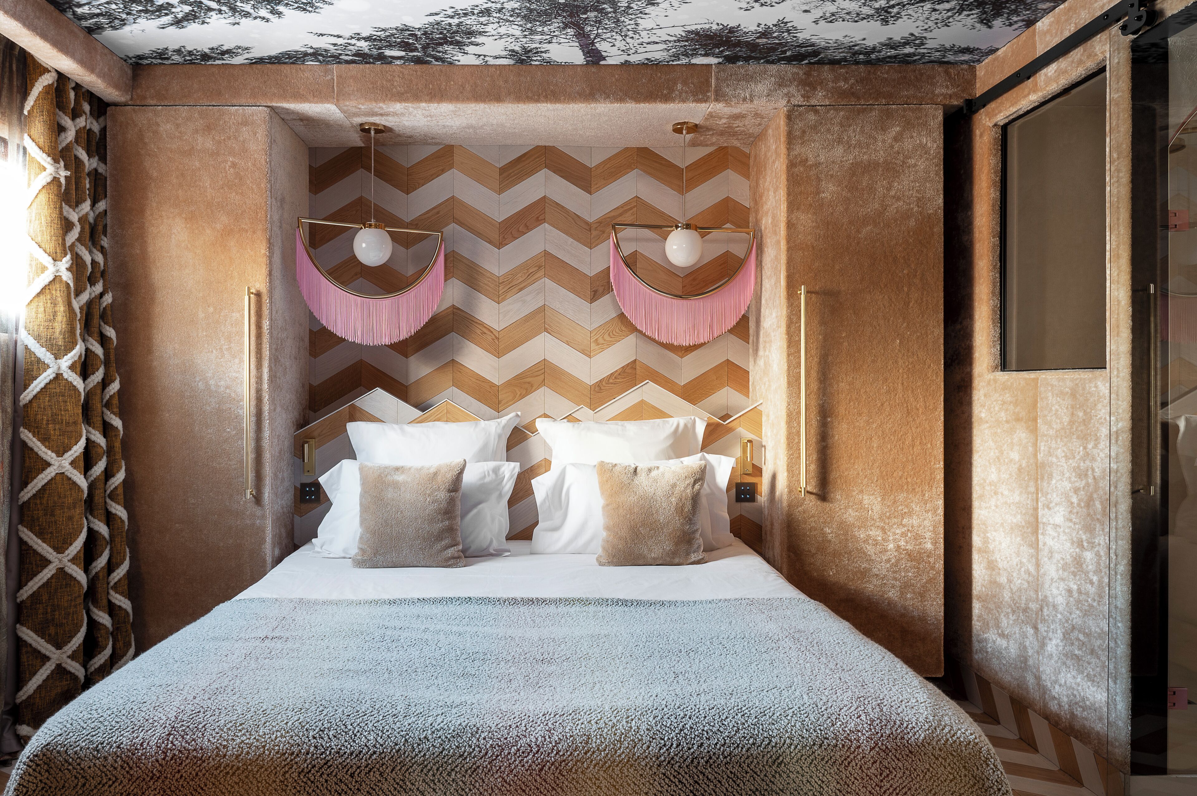 Une chambre avec un mur orné d’un motif en chevrons, un lit recouvert de linge de maison blanc et deux appliques murales roses à franges.