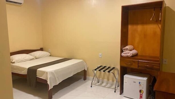 A room with a bed, a wooden wardrobe, a small refrigerator, and a radiator.