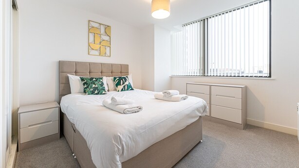 A neatly arranged bedroom with a large bed, a bedside table, a nightstand, a window with blinds, and a wall-mounted light fixture.
