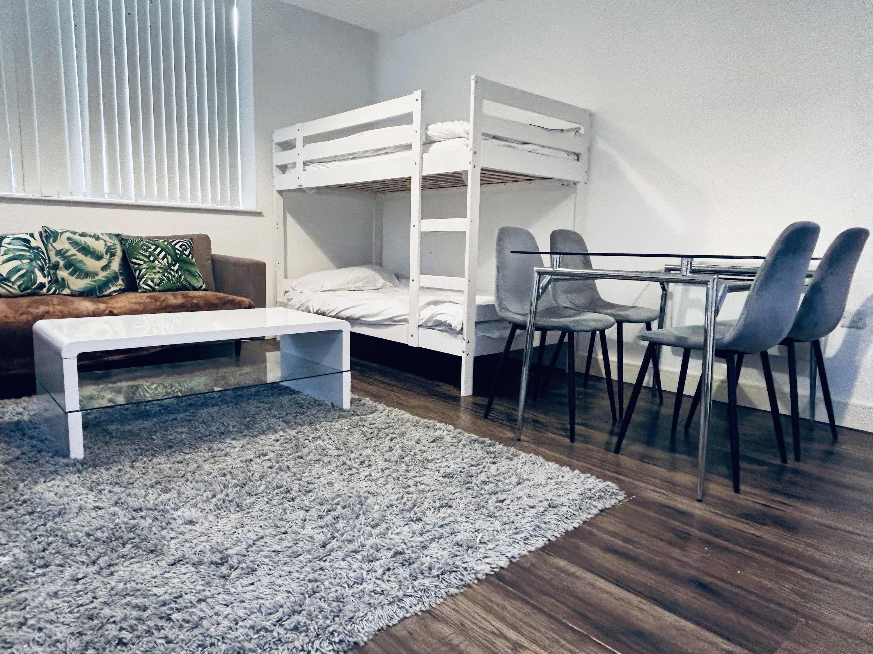 A bunk bed with a desk and chairs, a sofa, and a small table in a room with wooden flooring and a grey rug.