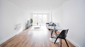 Un salón moderno y bien iluminado con una mesa redonda de cristal, sillas negras, un sofá gris y un ventanal que proporciona luz natural.