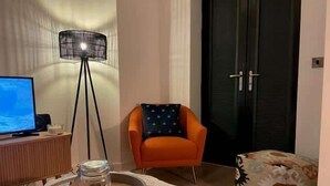 A modern living room with a television, a floor lamp, an orange armchair, a patterned ottoman, and a tray with a jar of nuts. - Central Solihull 1-Bed | Parking • WiFi • NEC (Solihull)