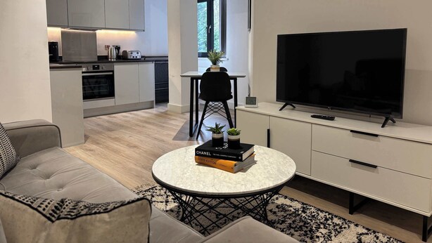 A modern living room with a sofa, a coffee table with books, a TV, and a kitchen in the background. - Solihull Studio • WiFi • NEC • BHX (Solihull)
