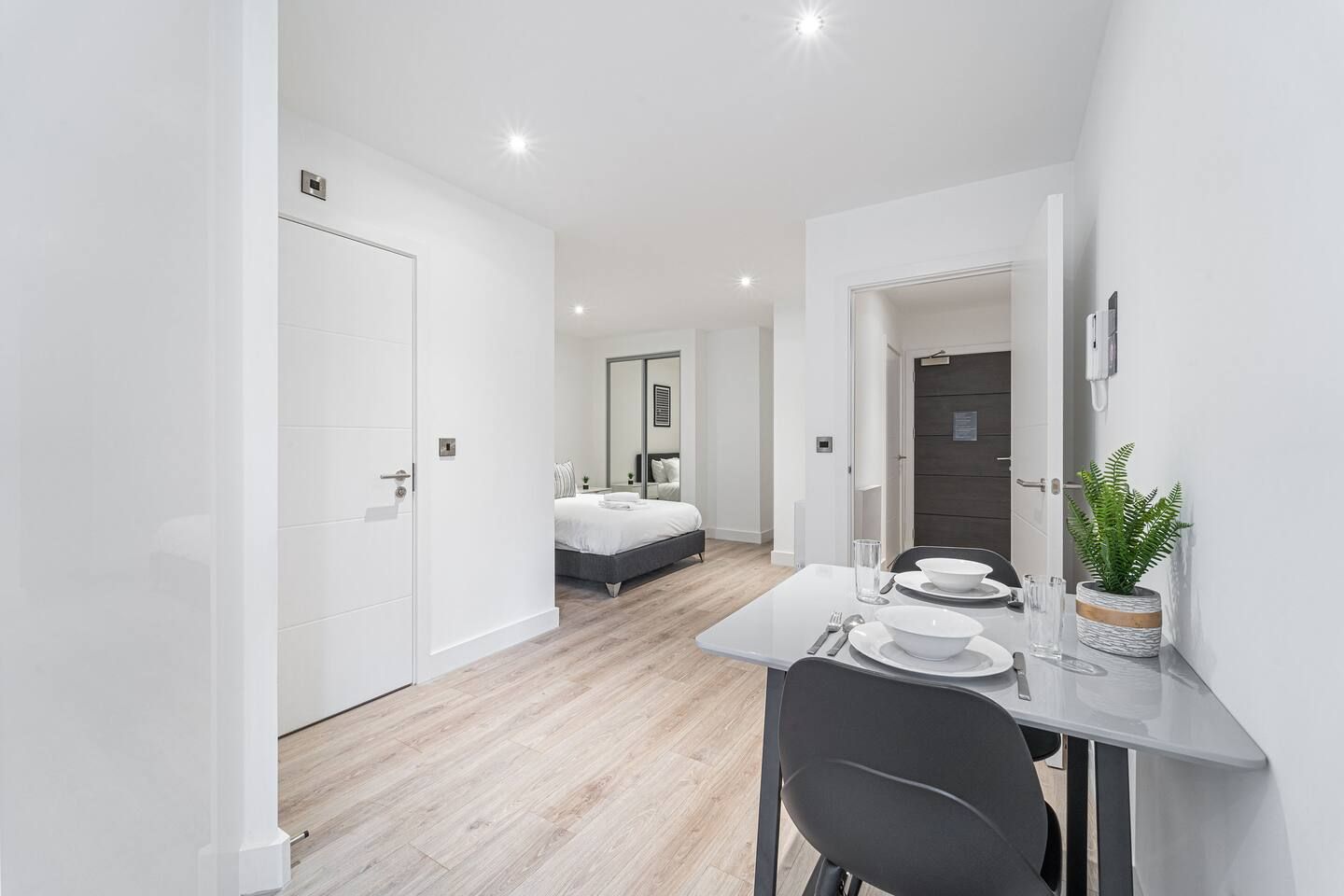 Une chambre moderne et minimaliste, comprenant un lit, un coin repas avec une table et des chaises, ainsi qu’une petite plante en pot.