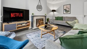 A modern living room with a flat-screen TV displaying 'NETFLIX', a fireplace, a sofa, a coffee table, and a floor lamp.