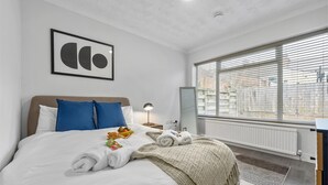 A hotel room with a bed, a bedside table with a lamp, a window with blinds, and a framed picture on the wall.