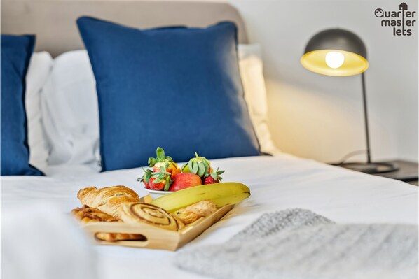 A neatly made bed with blue pillows, a bedside lamp, and a tray with pastries, fruit, and a banana.