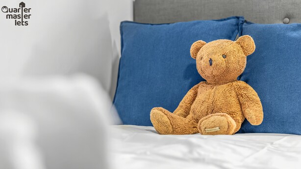 Un ours en peluche moelleux est assis sur un lit, à côté d’un oreiller bleu.