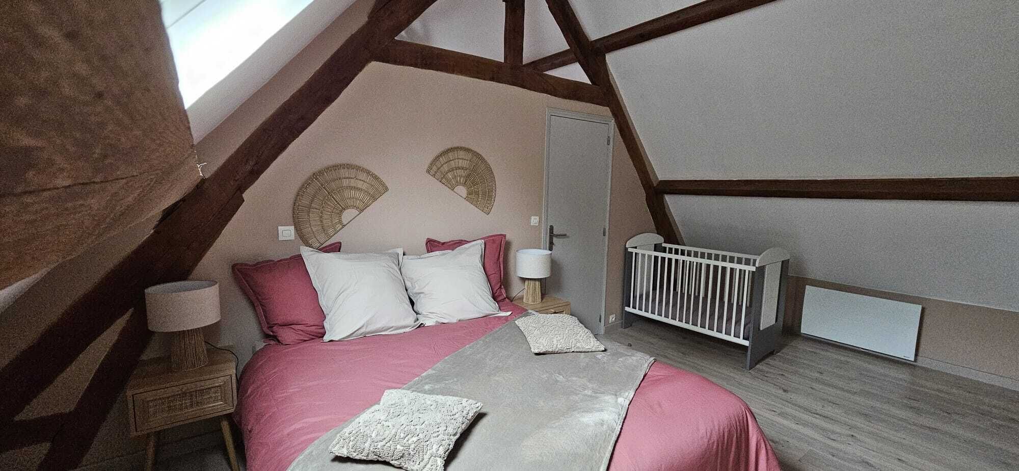 Un dormitorio con techo inclinado, cama con ropa de cama rosa, mesita de noche con lámpara y una cuna.