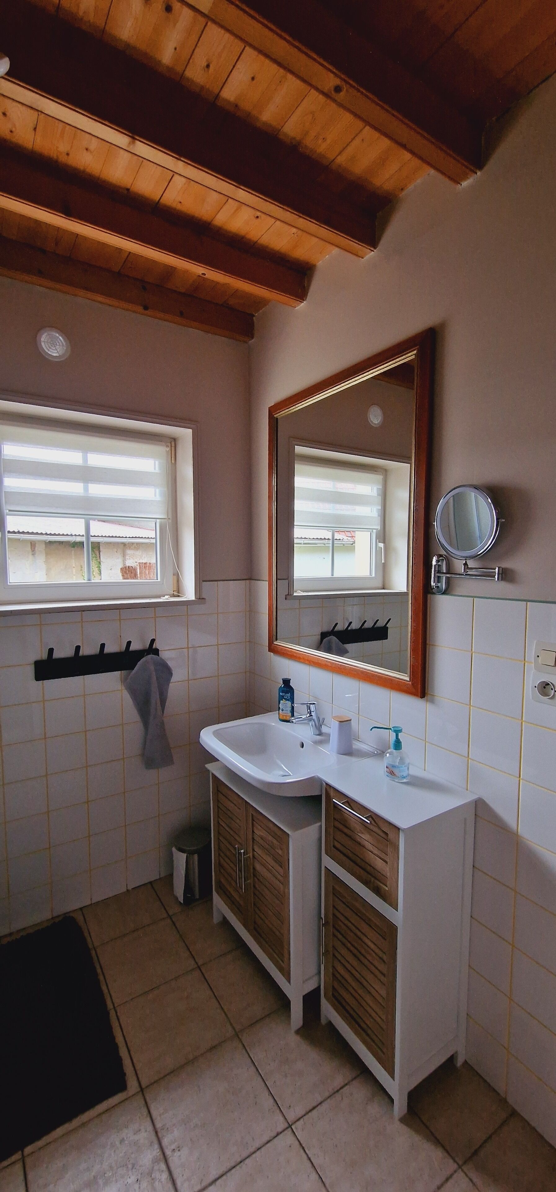 Une salle de bain avec un plafond en bois, un lavabo avec un miroir, un porte-serviettes et une fenêtre avec des stores.