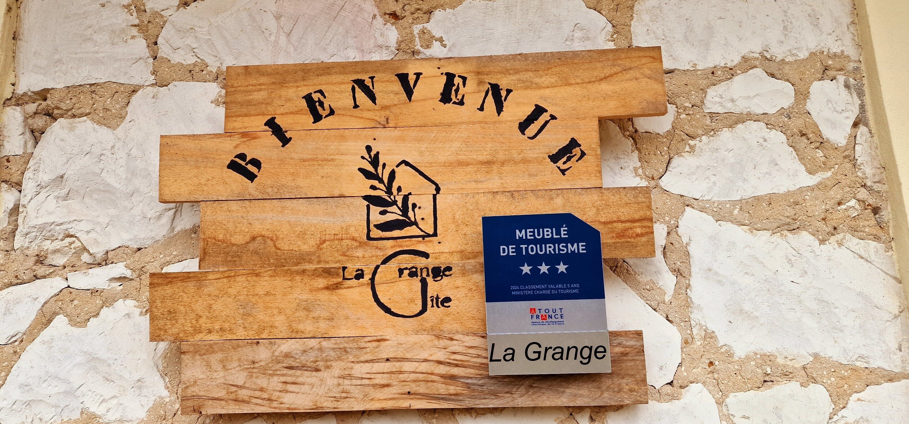 A wooden welcome sign with 'Bienvenue' and a house icon, a blue tourism award plaque, and 'La Grange' text.