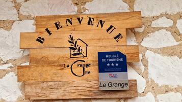 A wooden welcome sign with 'Bienvenue' and a house icon, a blue tourism award plaque, and 'La Grange' text.