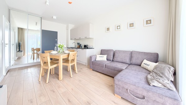 A modern living room with a grey sofa, wooden dining table, and chairs. There is a kitchen area with white cabinets and a microwave.