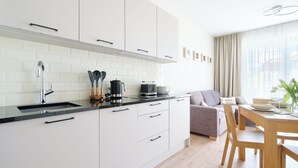 Une cuisine moderne avec des armoires blanches, un comptoir noir et des appareils en acier inoxydable. On y trouve un coin repas avec une table et des chaises, et un canapé à l’arrière-plan.