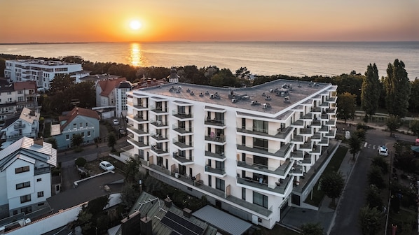 A modern multi-story building with balconies, located near a body of water with a sunset in the background.