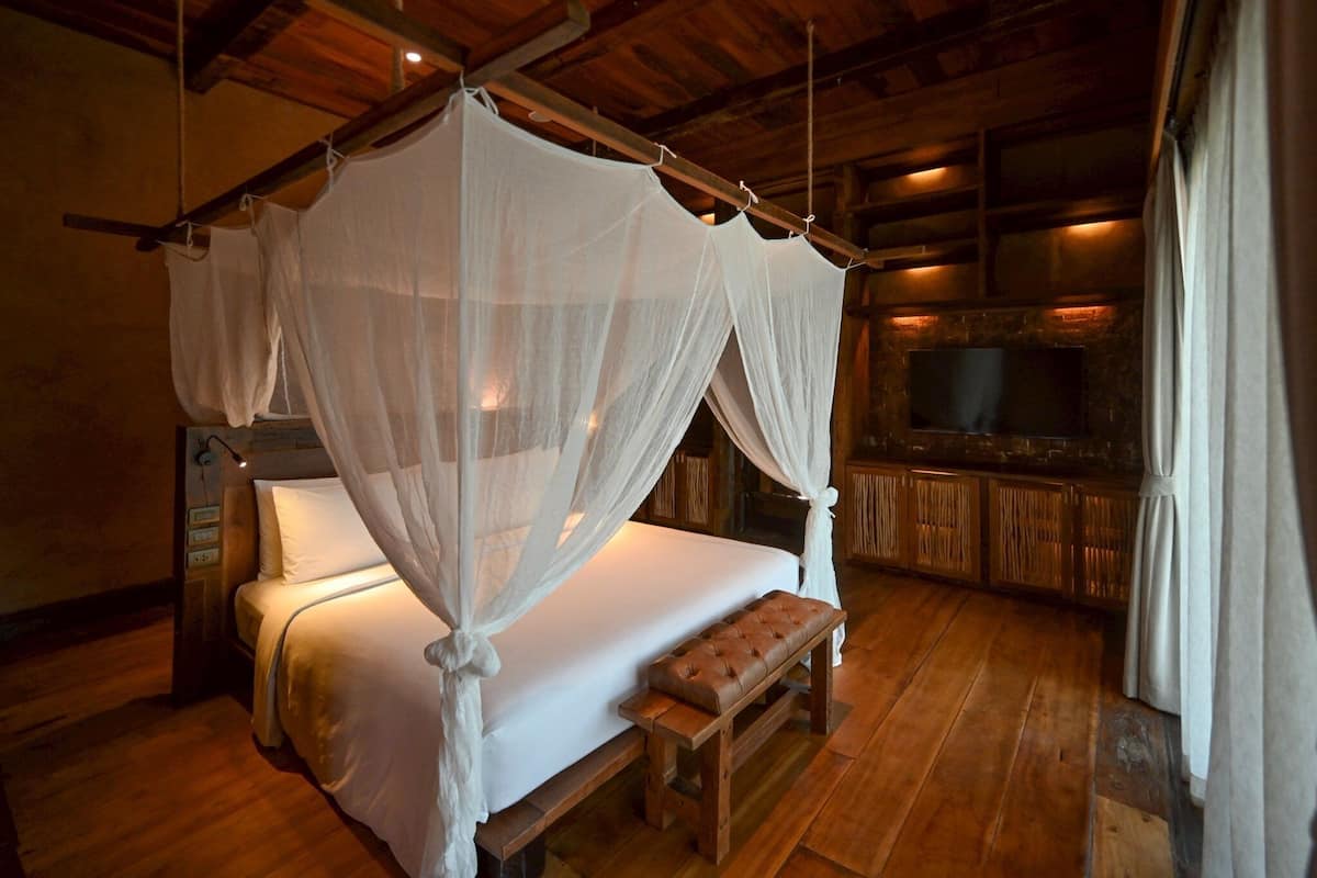 A four-poster bed with white linens and a bench, wooden flooring, and a stone wall with a television.