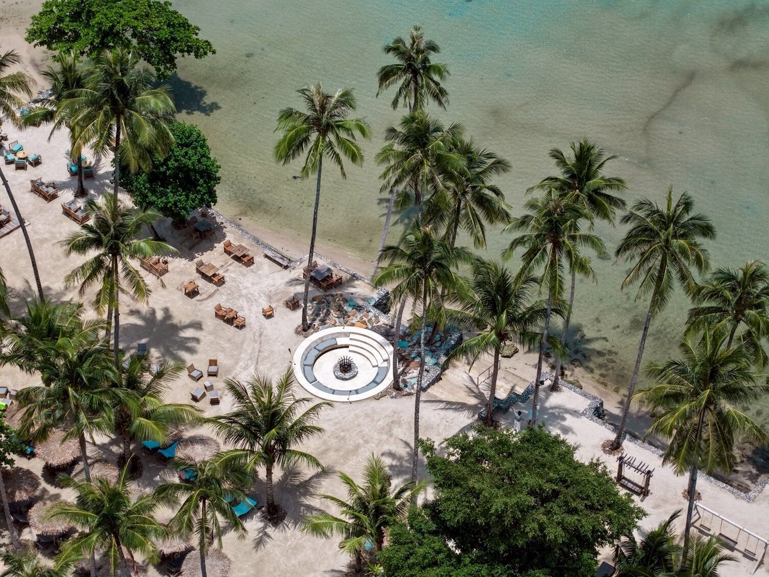 An aerial view of a beach with palm trees, lounge chairs, and a circular seating area.