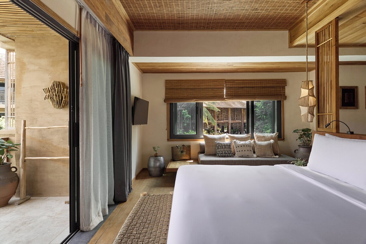 A bedroom with a large bed, a sofa, a TV, and a window with a view of greenery.