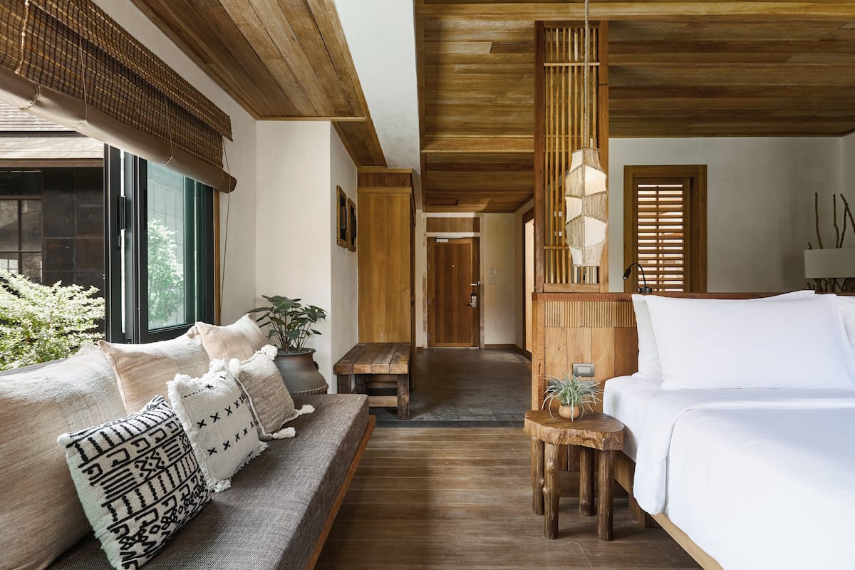 A room with a wooden ceiling, a bed with white bedding, a sofa with patterned cushions, and a view of greenery outside.