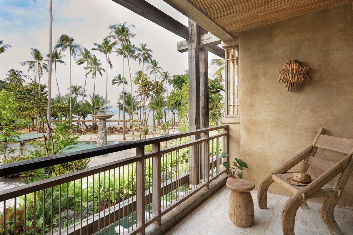 A balcony with a view of a pool and palm trees, wooden furniture, and a stone pillar.