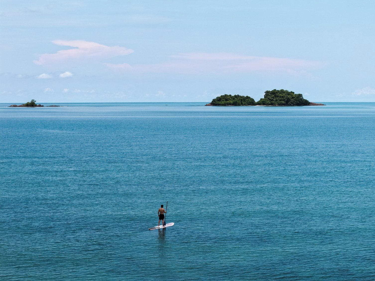 A person paddleboarding on calm blue waters with small islands in the distance.