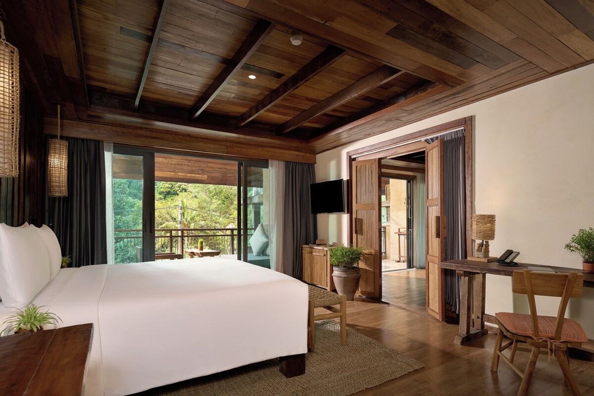 A spacious bedroom with a large bed, a desk, and a chair, a balcony with a view, and wooden ceiling beams.