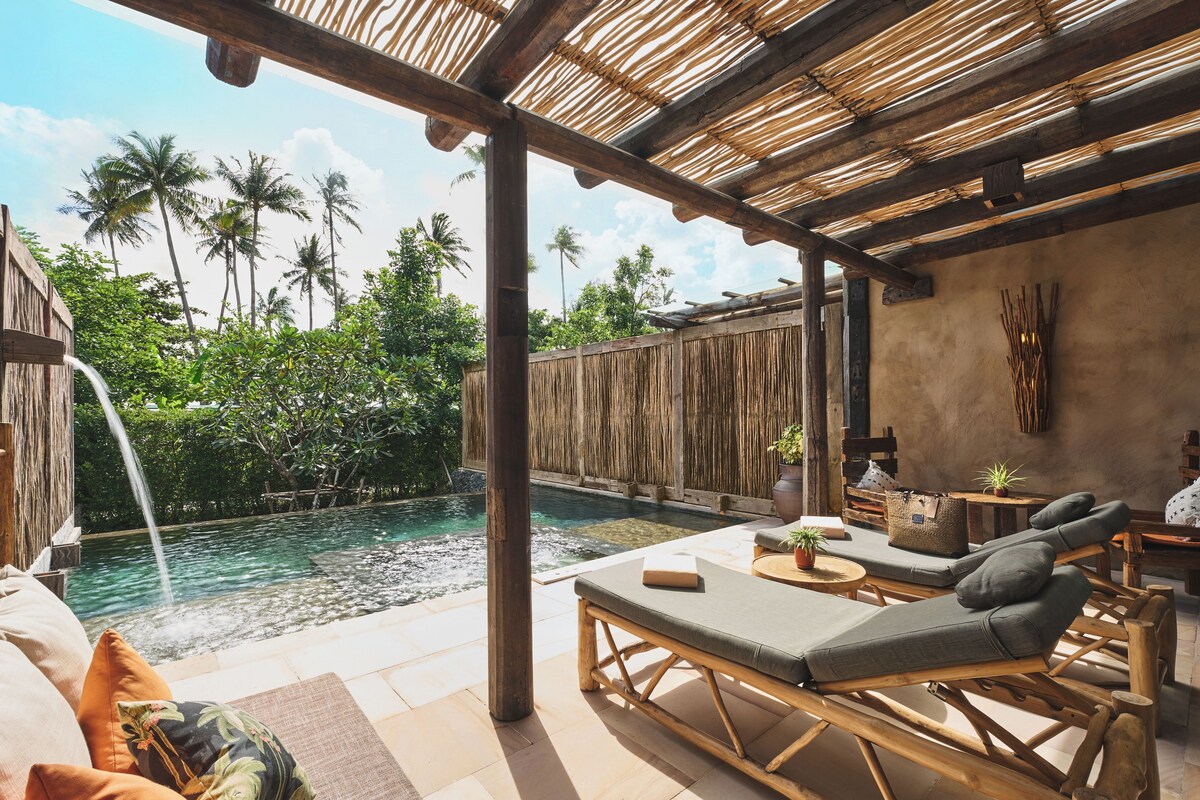 A poolside area with lounge chairs, a wooden pergola, and a view of a natural landscape with palm trees.