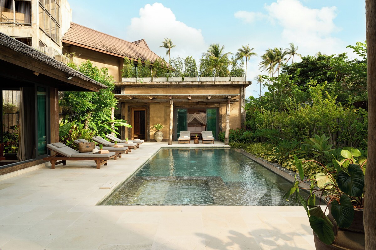 A swimming pool with lounge chairs and a covered seating area, surrounded by lush greenery and a building with a tiled roof.