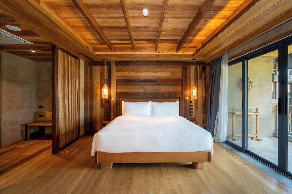 A wooden-paneled bedroom with a large bed, wooden floor, and a sliding glass door to a balcony.