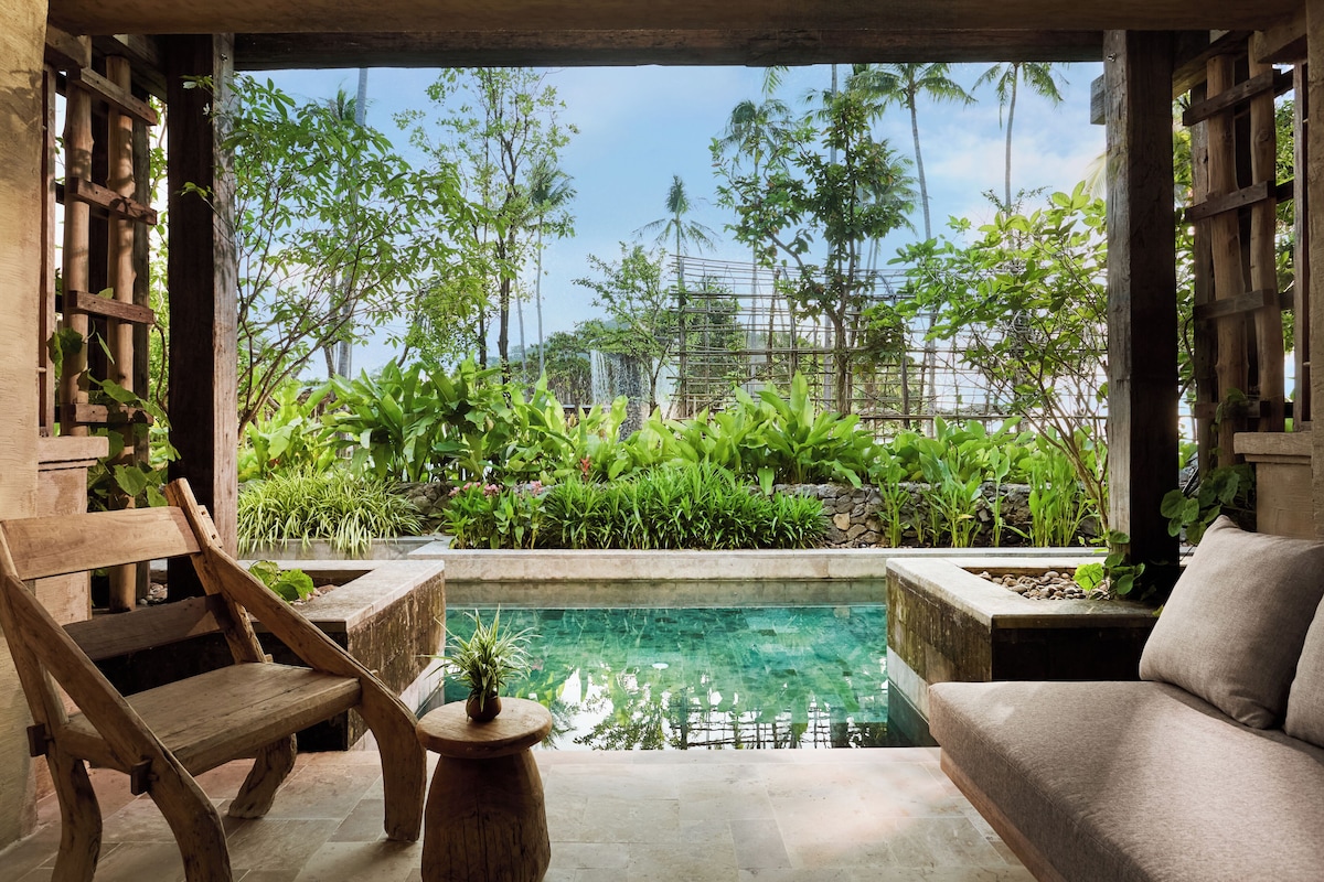 Eine Holzbank, ein kleiner Tisch mit Pflanze und eine Couch am Pool mit Blick auf Grünflächen und ein Gebäude im Hintergrund.