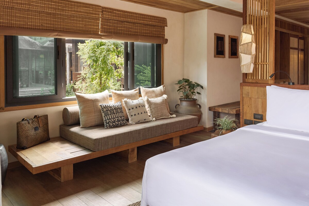 A bedroom with a large bed, a built-in bench with cushions, a window with a view of greenery, and a bedside table with a lamp.