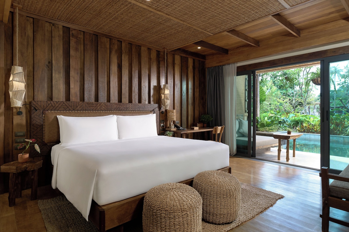 A bedroom with a large bed, wooden walls, a view of greenery outside, and a small table with a chair.