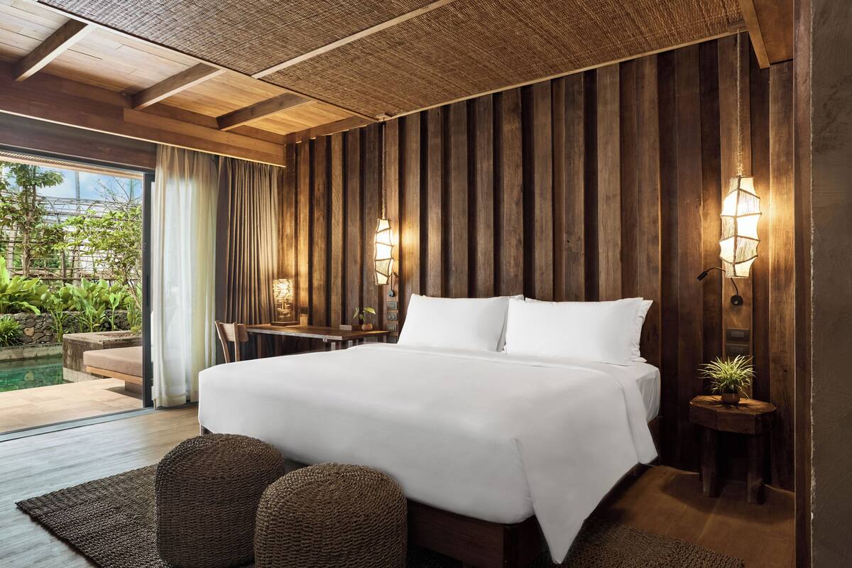A bedroom with a large bed, wooden panel walls, two bedside lamps, a small table, and a view of an outdoor pool area.