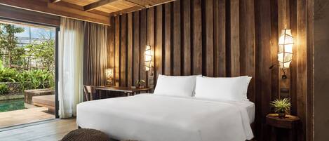 A bedroom with a large bed, wooden panel walls, two bedside lamps, a small table, and a view of an outdoor pool area.