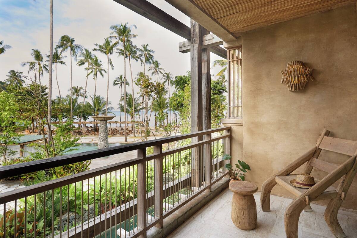 A balcony with a view of a pool and palm trees, wooden furniture, and a stone pillar.